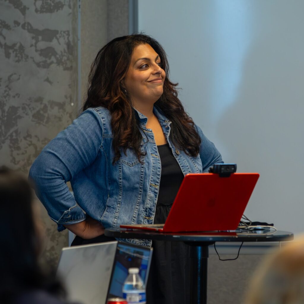 Woman standing at a podium with a laptop in a conference room while presenting, with audience members seated and informational posters in the background.